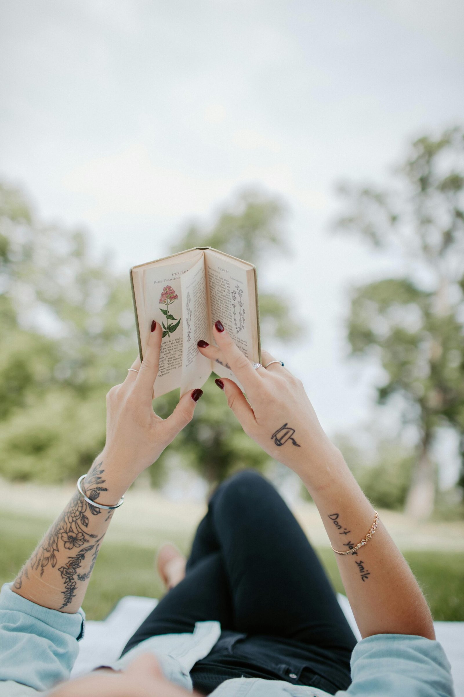 Person reading a book with tattoos visible, relaxing in a park on a sunny day.